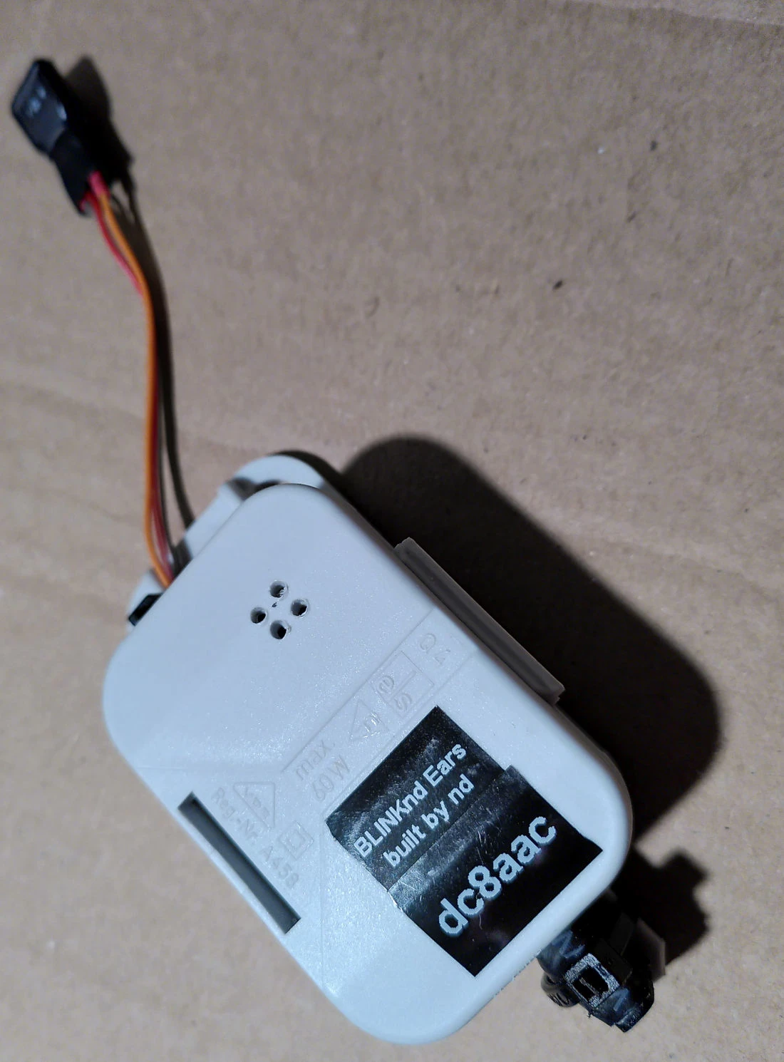 A white plastic box with a black label saying: 'BLINKnd Ears build by nd' and below that: 'dc8aac'. A cable with a plug comes out of the box, but isn#t attached to anything else.